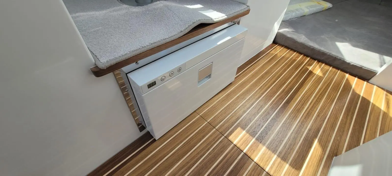 A white appliance on polished wooden flooring, beside a grey carpet and a light-coloured cushion, with controls visible.